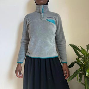 May include: A gray fleece pullover with a high collar and a teal accent pocket. The pullover has teal trim on the collar, cuffs, and hem. A small logo is visible on the teal pocket. The garment is part of a casual clothing collection.