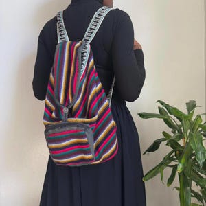 Puede incluir: Mochila a rayas con rayas verticales rojas, moradas, amarillas y marrones. Tiene un pequeño bolsillo delantero y correas blancas con ribete decorativo. La mochila está hecha de tela tejida.