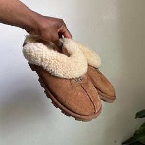 May include: A pair of tan suede slippers featuring a thick, cream-colored sheepskin lining and collar. The slippers have a rounded toe, a visible center seam, and a thick, textured rubber sole. The UGG brand logo is embossed on the front.