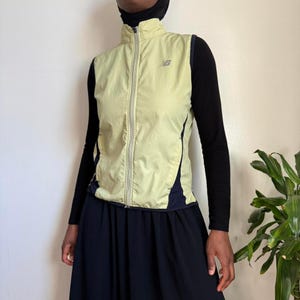 May include: A pale yellow New Balance vest with a full zip and a small logo on the chest. The vest features navy blue side panels and a high collar. The vest is worn over a black long-sleeved top and a black skirt.