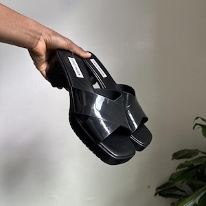 May include: Black platform sandals with a glossy finish. The sandals feature a thick sole and a crisscross strap design. The brand name "STEVE MADDEN" is visible on the insole. The shoes are held by a hand.