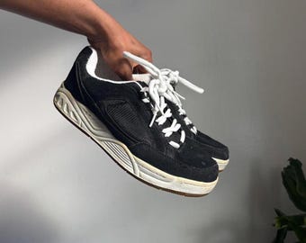 Pode incluir: Sapatos esportivos pretos e brancos com cadarços brancos. Os sapatos têm cabedal de camurça e sola grossa branca suja. Os sapatos são segurados por uma mão.