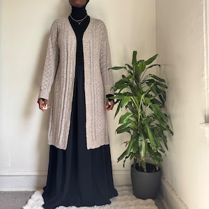 May include: A long, open-front beige cardigan featuring a textured knit pattern with vertical cable-knit details. The garment is worn over a black long-sleeved top and a long, flowing black skirt, creating a layered look with neutral tones.