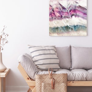 May include: A light gray sofa with two pillows, one with white and black stripes, and a woven basket with leather handles. The sofa is in front of a white wall with a framed abstract painting with purple, pink, and blue colors.