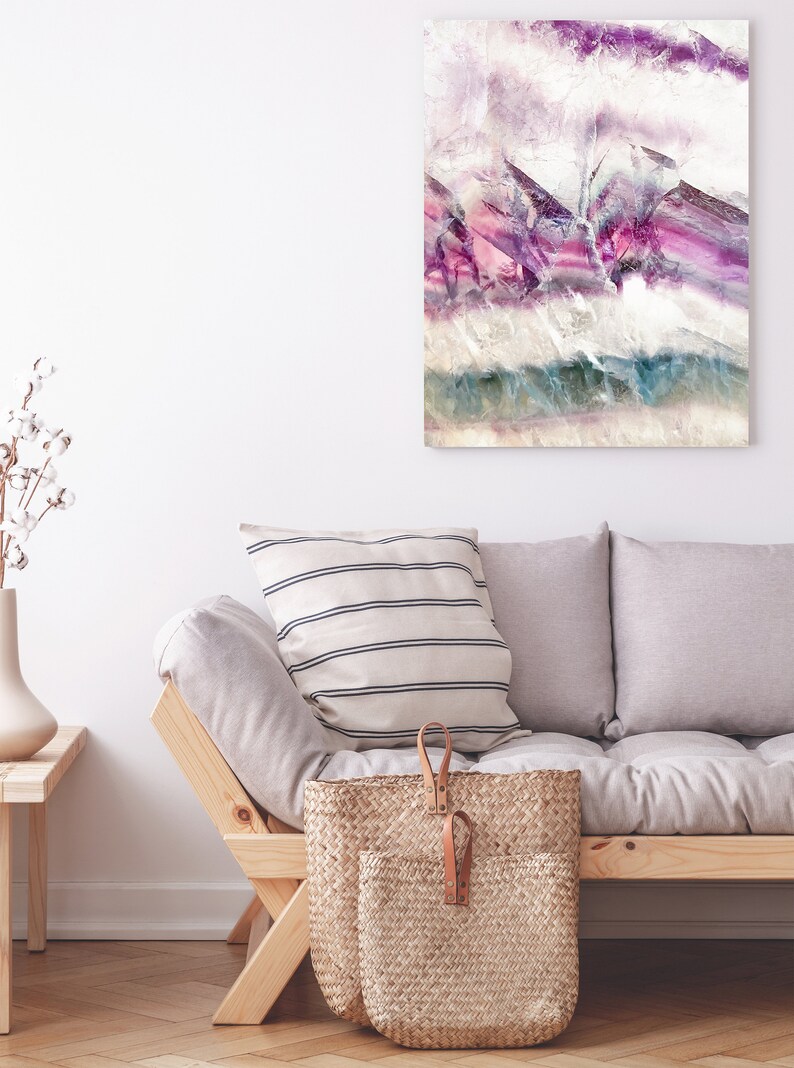 May include: A light gray sofa with two pillows, one with white and black stripes, and a woven basket with leather handles. The sofa is in front of a white wall with a framed abstract painting with purple, pink, and blue colors.