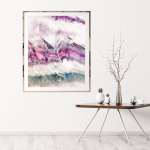 May include: Abstract art print in a gold frame with purple, pink, and blue hues. The print is hanging on a white wall above a wooden table with a vase and decorative items.