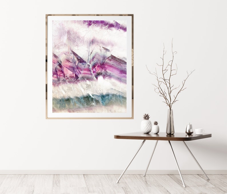 May include: Abstract art print in a gold frame with purple, pink, and blue hues. The print is hanging on a white wall above a wooden table with a vase and decorative items.