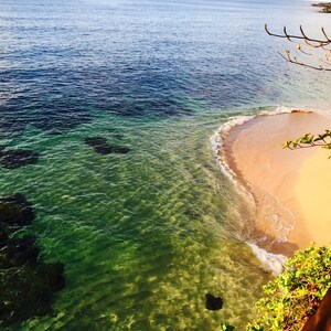 May include: A view from a cliff overlooking a small, sandy beach with clear, turquoise water. The water is shallow and you can see the ocean floor.