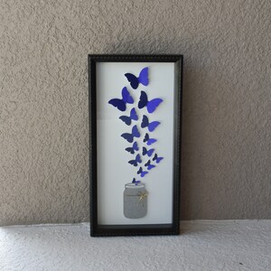 May include: A framed artwork featuring a jar with butterflies. The butterflies, in shades of blue, appear to be emerging from the jar. The artwork is in a black rectangular frame, creating a visually appealing piece.