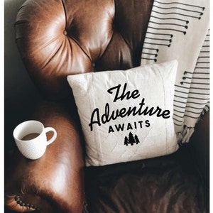 May include: A white throw pillow with black text that reads "The Adventure Awaits" and a small graphic of three pine trees. The pillow is sitting on a brown leather armchair with a white and black striped throw blanket.