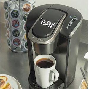 Op de afbeelding: Een zwarte Keurig koffiezetapparaat met een zilveren bovenkant en een witte mok gevuld met koffie. Het koffiezetapparaat heeft een digitaal display en een knop om verschillende brouwmaten te selecteren. De bovenkant van het koffiezetapparaat heeft een sticker met de tekst "But First Coffee".