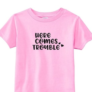 Op de afbeelding: Roze T-shirt met korte mouwen met zwarte tekst die "Here comes trouble" zegt met twee kleine hartjes.