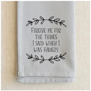 May include: A gray kitchen towel with black text that reads "Forgive me for the things I said when I was hangry". The text is framed by a simple black floral design.