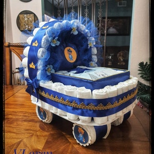 Puede incluir: Un pastel de pañales azul y dorado con forma de cochecito. El cochecito está decorado con cinta azul y dorada, tul y flores. El pastel está coronado con una corona azul y dorada y un pequeño animal de peluche.