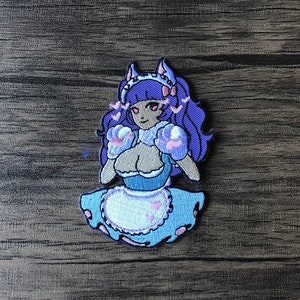 May include: An embroidered patch featuring a cartoon character with purple hair, wearing a blue and white maid outfit with pink accents. The character has cat ears and is holding pink boxing gloves.