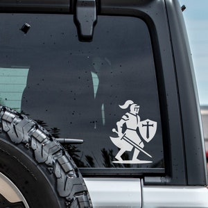 Op de afbeelding: Witte vinyl sticker van een ridder in harnas met een zwaard en schild met een kruis erop. De sticker zit op de achterruit van een witte auto.