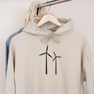 Puede incluir: Una sudadera con capucha beige con un gr&aacute;fico de turbina e&oacute;lica negra en la parte delantera. La sudadera est&aacute; colgada de una percha de madera.