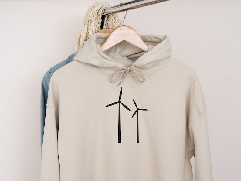 Puede incluir: Una sudadera con capucha beige con un gr&aacute;fico de turbina e&oacute;lica negra en la parte delantera. La sudadera est&aacute; colgada de una percha de madera.