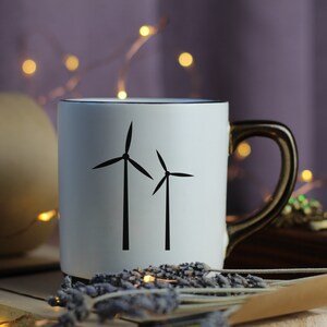 Puede incluir: Taza de cer&aacute;mica blanca con un contorno negro y una silueta negra de dos aerogeneradores. La taza tiene un asa dorada.