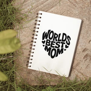 K&ouml;nnte beinhalten: Ein wei&szlig;es Notizbuch mit einer schwarzen Spiralbindung. Das Notizbuch tr&auml;gt die Worte "Worlds Best Mom" in Herzform.