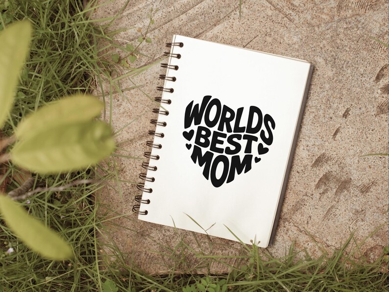 K&ouml;nnte beinhalten: Ein wei&szlig;es Notizbuch mit einer schwarzen Spiralbindung. Das Notizbuch tr&auml;gt die Worte "Worlds Best Mom" in Herzform.