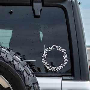 May include: A white wreath sticker on the back window of a black car. The wreath is made of simple leaves and berries.