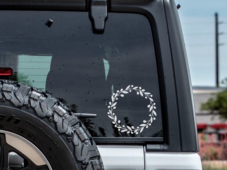 May include: A white wreath sticker on the back window of a black car. The wreath is made of simple leaves and berries.