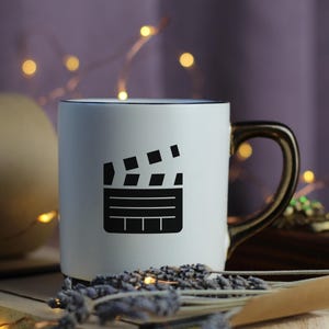 May include: White ceramic mug with a black outline and a black and white image of a clapperboard. The mug has a gold handle.