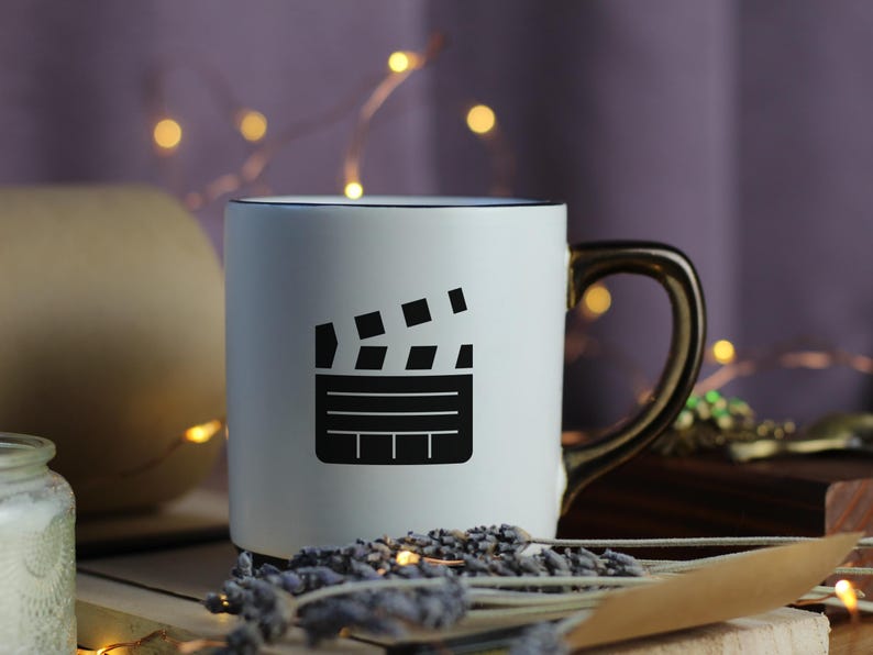May include: White ceramic mug with a black outline and a black and white image of a clapperboard. The mug has a gold handle.