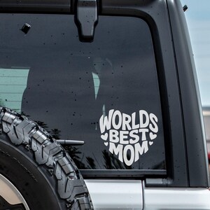 K&ouml;nnte beinhalten: Ein wei&szlig;er Vinyl-Aufkleber mit dem Text "Worlds Best Mom" in Herzform, angebracht an der Heckscheibe eines silbernen Autos.