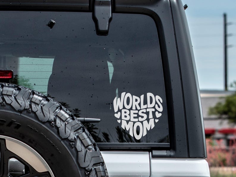 K&ouml;nnte beinhalten: Ein wei&szlig;er Vinyl-Aufkleber mit dem Text "Worlds Best Mom" in Herzform, angebracht an der Heckscheibe eines silbernen Autos.