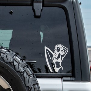 Op de afbeelding: Witte vinyl sticker van een vrouw met lang haar die een surfboard vasthoudt. De sticker zit op de achterruit van een auto.