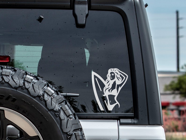 Op de afbeelding: Witte vinyl sticker van een vrouw met lang haar die een surfboard vasthoudt. De sticker zit op de achterruit van een auto.