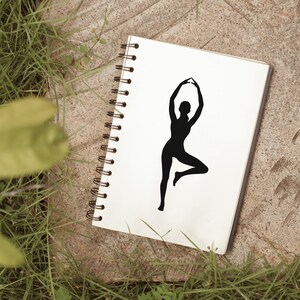Peut inclure: Un carnet blanc avec une silhouette noire d'une personne en position de yoga sur la couverture. Le carnet est pos&eacute; sur une surface brune avec de l'herbe verte en arri&egrave;re-plan.