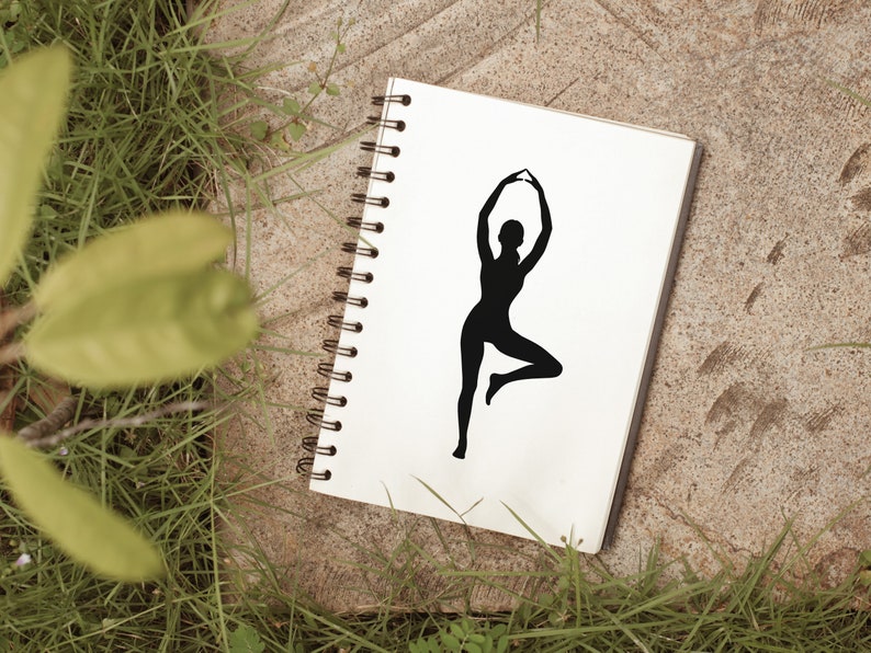 Peut inclure: Un carnet blanc avec une silhouette noire d'une personne en position de yoga sur la couverture. Le carnet est pos&eacute; sur une surface brune avec de l'herbe verte en arri&egrave;re-plan.