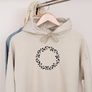 May include: A beige hoodie with a black floral wreath design on the front. The hoodie has a drawstring hood and is hanging on a hanger.