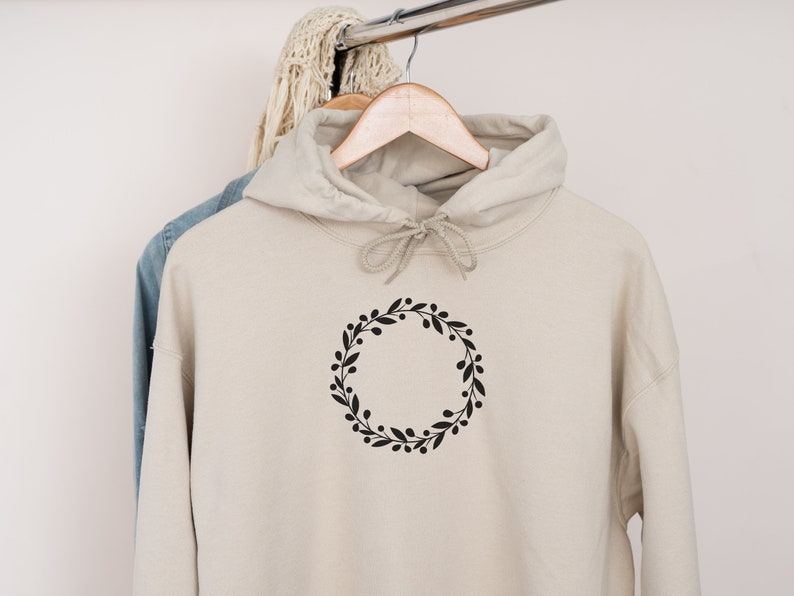 May include: A beige hoodie with a black floral wreath design on the front. The hoodie has a drawstring hood and is hanging on a hanger.