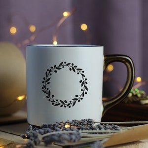 May include: White ceramic mug with a black wreath design. The mug has a gold handle and is sitting on a wooden surface with dried lavender.
