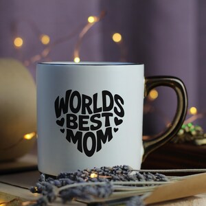 K&ouml;nnte beinhalten: Wei&szlig;e Keramiktasse mit einem schwarzen Herzdesign, auf dem "Worlds Best Mom" steht. Die Tasse hat einen schwarzen Rand und einen goldenen Henkel.