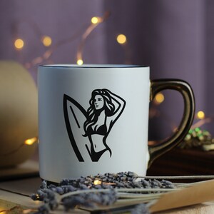 Op de afbeelding: Een witte keramische mok met een zwarte omtrek en een zwart-witte afbeelding van een vrouw die een surfboard vasthoudt. De mok heeft een gouden handvat.