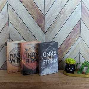 May include: Three paperback books with fantasy covers. The books are titled "Four Winds", "Iron Flame", and "Onyx & Storm". The author is Rebecca Yarros. The books are on a wooden surface with two small potted plants.