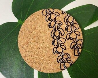 May include: A round cork coaster with a black floral design. The coaster is on a green leaf.