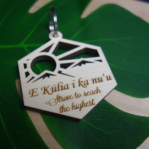 May include: Wooden keychain with a geometric design featuring a sun, mountains, and the text "E Kūlia i ka nu'u, Strive to reach the highest." The keychain is light brown with a silver keyring.