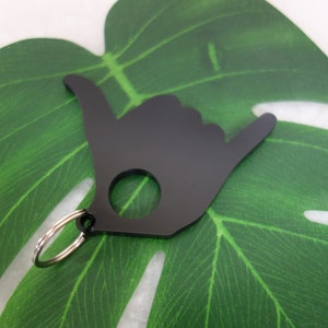 May include: Black metal keychain shaped like a hand giving the shaka sign. The keychain has a silver ring for attaching keys.