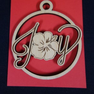 May include: A wooden ornament shaped like a circle with the word "Joy" written in a cursive font. A white hibiscus flower is centered in the word "Joy". The ornament is on a red background.