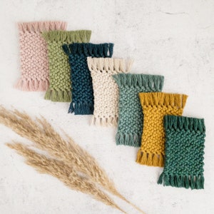 May include: A set of seven woven coasters in various colors, including pink, green, blue, beige, and yellow. The coasters have a fringed edge and are arranged in a row on a white surface.