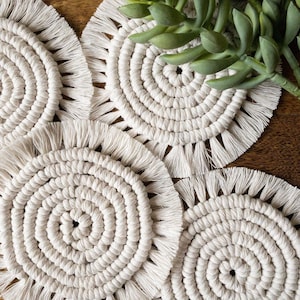 May include: Set of four white crocheted coasters with fringe detailing. The coasters are circular and have a woven pattern.