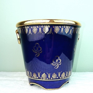 Może przedstawiać: Kobaltowo-niebieski ceramiczny pojemnik na lód ze złotymi wykończeniami i dekoracyjnymi złotymi wzorami kwiatowymi i geometrycznymi. Pojemnik ma dwa małe uchwyty i złotą obręcz. Podstawa ma trzy małe nóżki.