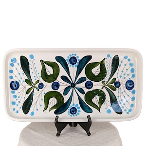 Pode incluir: Uma bandeja retangular de cerâmica branca com um padrão botânico simétrico pintado à mão em tons de verde escuro, azul-petróleo e azul claro. O design inclui folhas estilizadas, caules finos e pequenos detalhes florais circulares.
