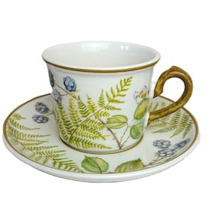May include: A white teacup and saucer set with a botanical design. The cup and saucer feature a fern and blackberry pattern in green, blue, and white. The cup has a brown handle and trim. The saucer is round.